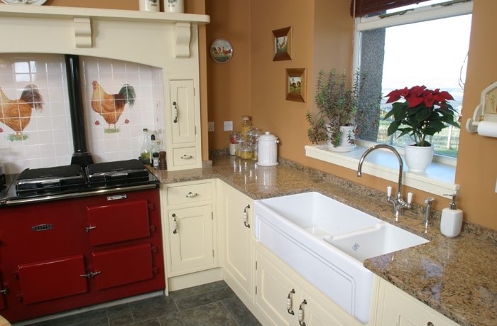 Gemütliche Landhausküche mit cremefarbenen Schränken, rotem Herd und nostalgischer Keramikspüle Landhausküche mit hellen Einbauschränken, rotem Herd, weißen Fliesen mit Hühner-Motiv, Granit-Arbeitsplatte, Keramikspüle und Fensterbank mit Zimmerpflanzen