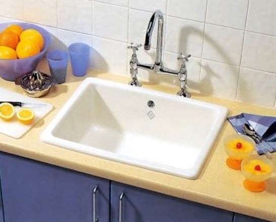 Küchenspüle aus Keramik mit Doppelarmatur und frischem Obst auf Arbeitsplatte Weiße Keramik-Küchenspüle auf beiger Arbeitsplatte mit Doppelarmatur, umgeben von Orangen, Gläsern, Zitronenhälften und Küchenutensilien