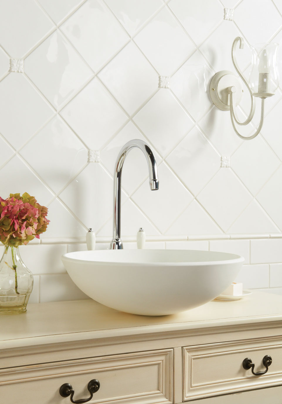 Waschbecken im Landhausstil mit Chromarmatur und dekorativer Retro-Wandleuchte Modernes, weißes Aufsatzwaschbecken mit Chromarmatur auf nostalgischer Kommode, dahinter weiße Wandfliesen im Landhausstil, seitlich eine Glasvase mit rosafarbener Blume und eine dekorative Wandleuchte