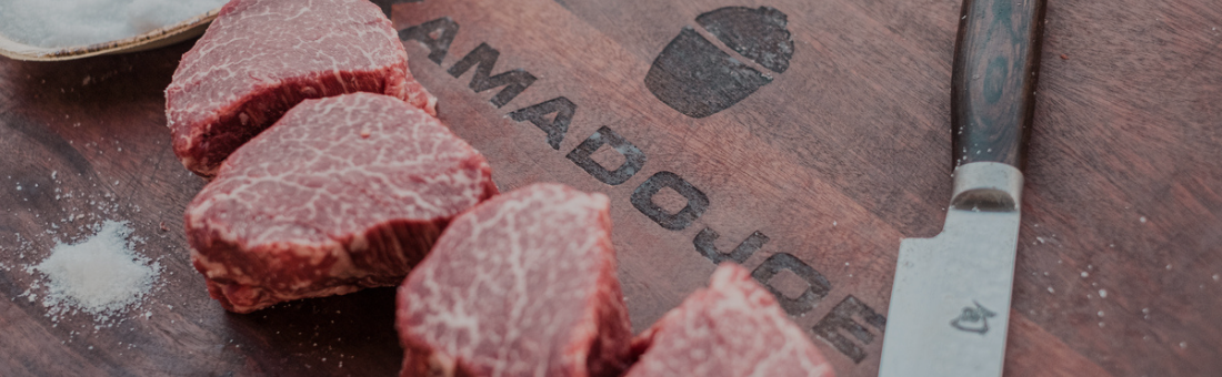 Rohe Steaks auf Kamado Joe Schneidebrett mit Messer und Salz Mehrere rohe Rindersteaks liegen auf einem Holzschneidebrett von Kamado Joe, daneben ein Messer und etwas Salz