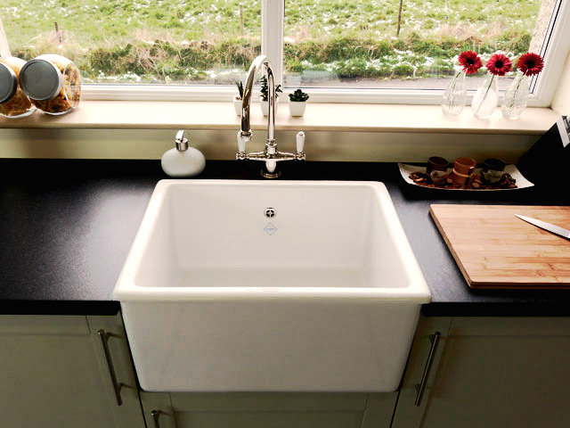 Moderne Küchenspüle aus Keramik mit Armatur vor Fenster, schwarze Arbeitsplatte Weiße, eckige Keramik-Küchenspüle mit stilvoller Armatur, darunter dunkle Arbeitsplatte, vor einem Fenster mit Ausblick und Dekoration