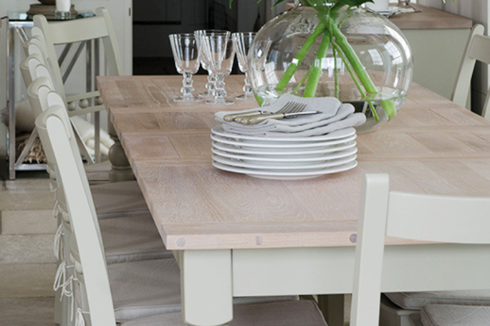 Heller Esstisch aus Holz mit Stühlen, gestapelten weißen Tellern, Gläsern und einer Vase mit frischen Blumen als Tischdekoration Detailaufnahme eines gedeckten Esstisches aus Holz mit weißen Stühlen, gestapelten Tellern, Besteck, Gläsern und einer Glasvase mit grünen Zweigen