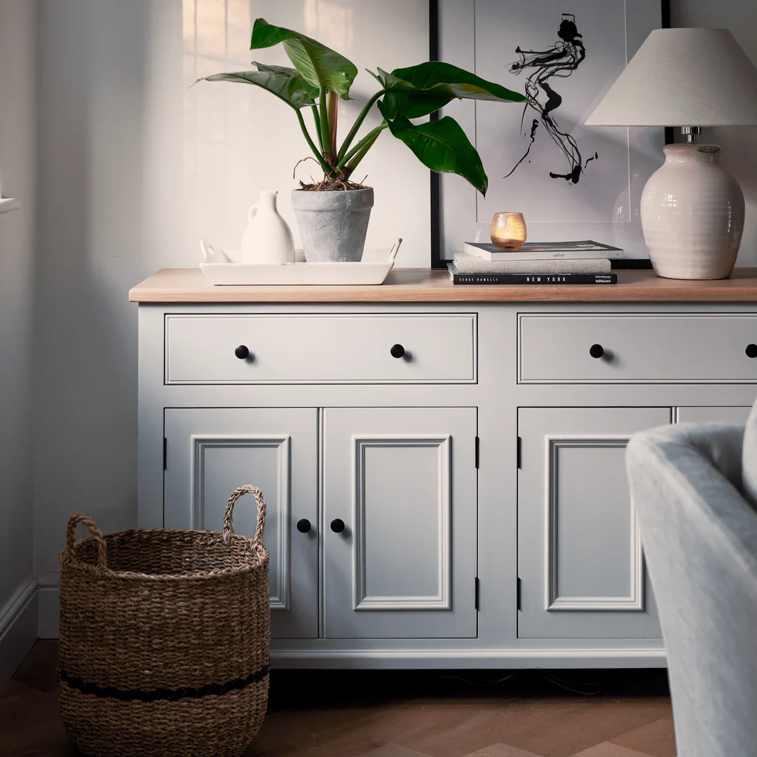 Helles Sideboard im Landhausstil in Grau mit Holzplatte, 2 Schubladen und 4 Türen; dekoriert mit Zimmerpflanze, Keramiklampe und Tablett; Einsatz im Wohnzimmer mit Korb