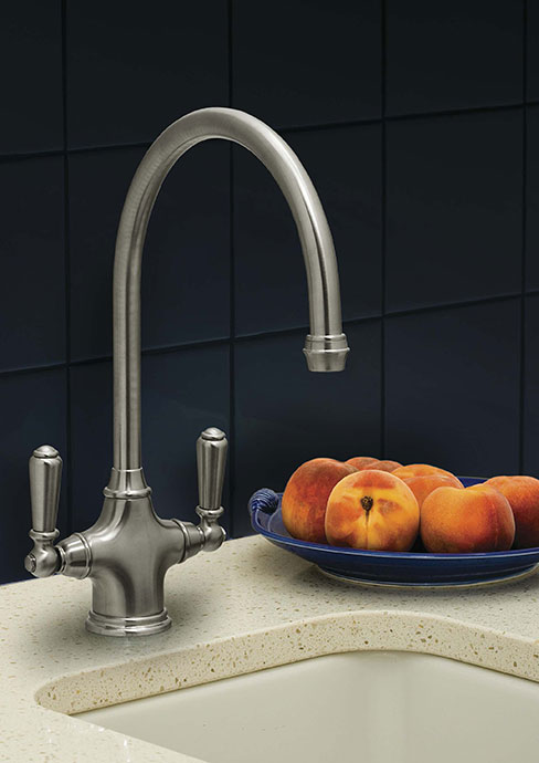 Edelstahl-Küchenarmatur mit zwei Griffen neben einer Schale mit frischen Pfirsichen auf Küchenarbeitsplatte Moderne Küchenarmatur aus Edelstahl mit zwei Griffen, daneben steht auf der Arbeitsplatte eine blaue Schale mit Pfirsichen