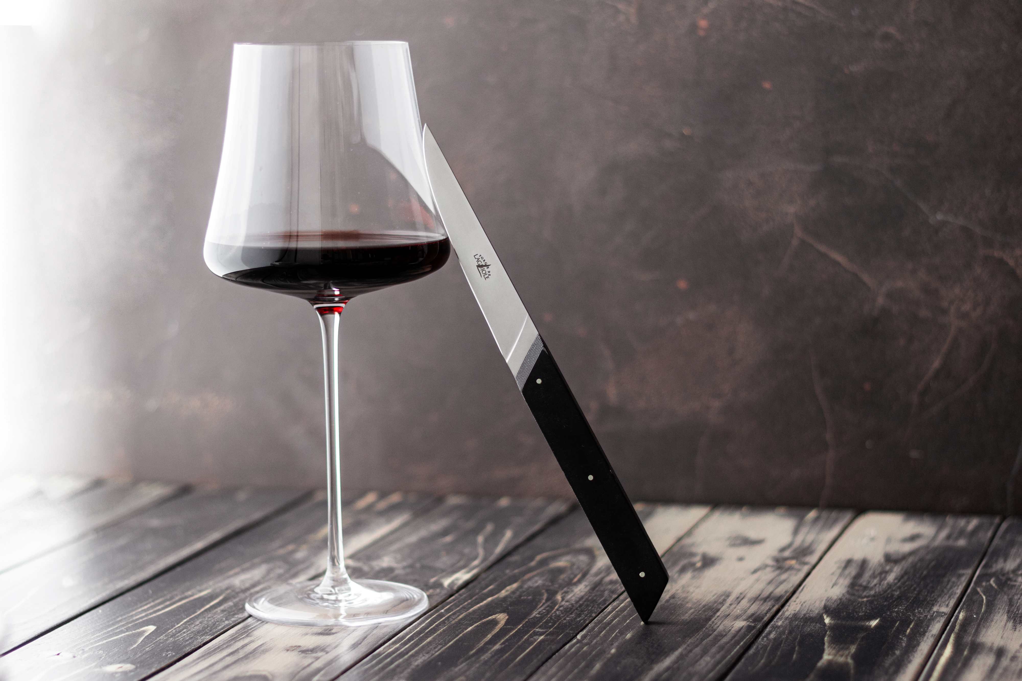 Weinglas mit Rotwein neben einem schwarzen Messer auf einem dunklen Holztisch vor rustikalem Hintergrund