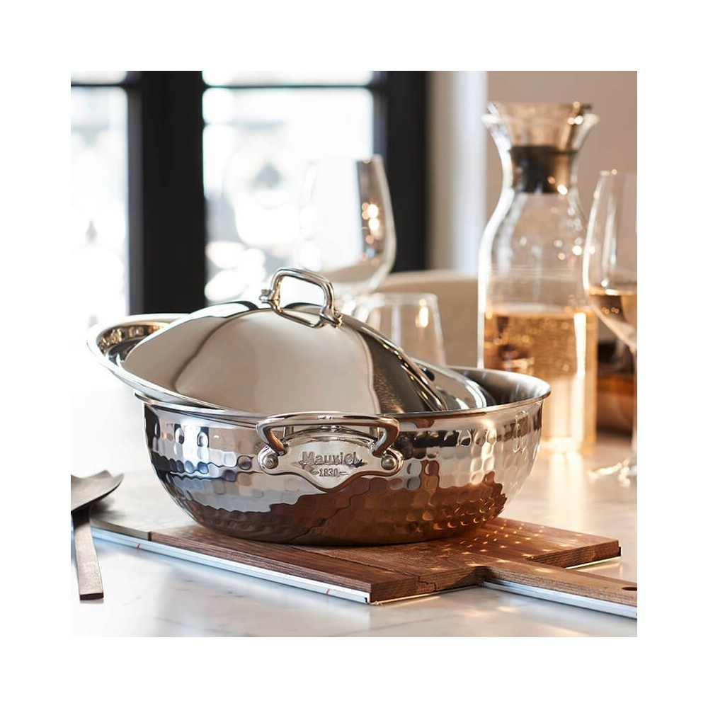 Eleganter Kupfertopf mit Hammerschlag-Optik und Deckel von Mauviel auf einem gedeckten Tisch Hochwertiger Kupfertopf mit gehämmerter Oberfläche und Deckel, platziert auf einem Holzbrett, umgeben von Gläsern und einer Karaffe auf einem Esstisch