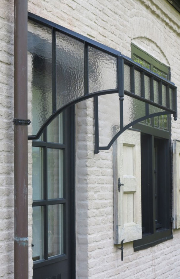 Moderne Haustür Überdachung aus Glas mit schwarzem Metallrahmen für stilvolle Fassaden Glasüberdachung mit schwarzem Metallrahmen über einer Haustür an einer hellen Ziegelhausfassade mit Fensterläden
