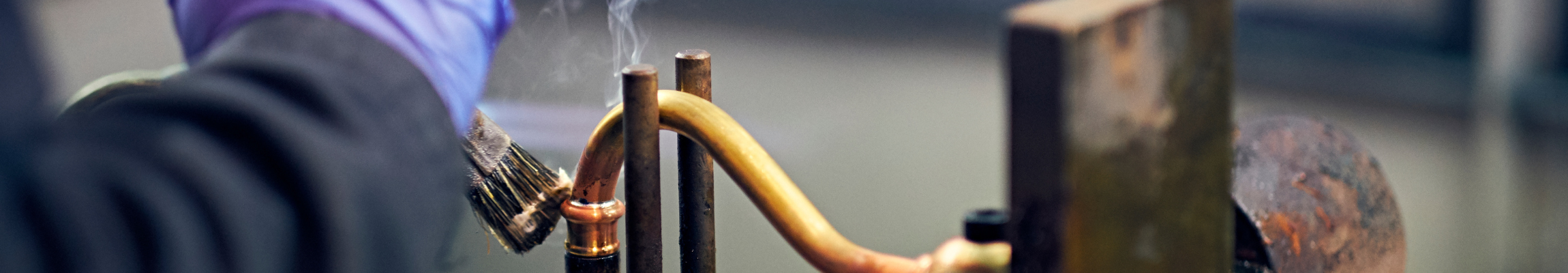 Handwerkliche Lötarbeiten an Kupferrohren – präzises Verbinden von Metallteilen Eine Person führt mit Handschuhen Lötarbeiten an einem gebogenen Kupferrohr durch. Es steigt Rauch auf.