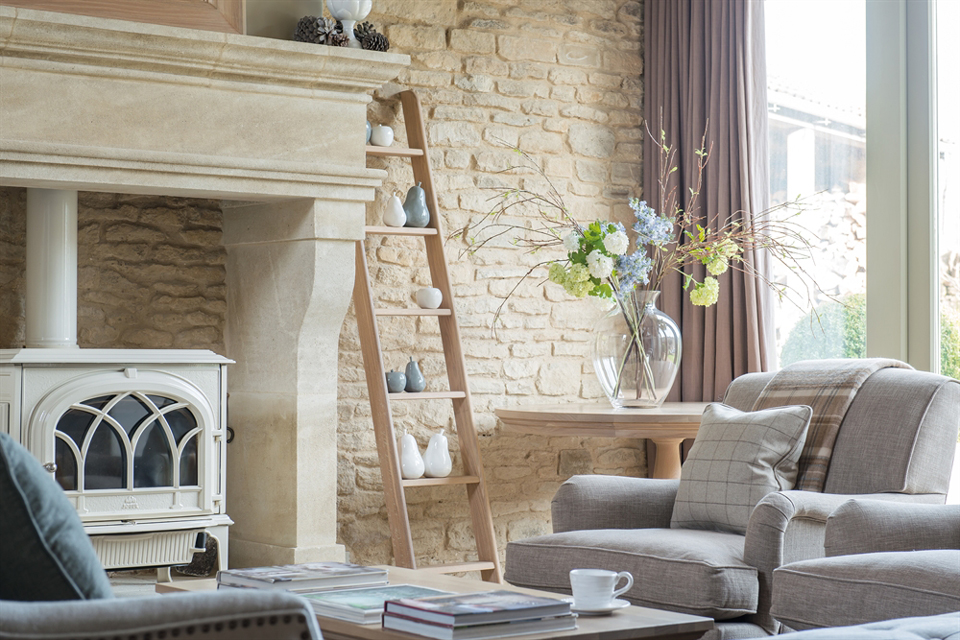 Gemütliches Wohnzimmer mit Kamin, Sprossenleiter, Dekoration und bequemen Polstermöbeln im Landhausstil Helles Wohnzimmer mit Kamin, Deko auf Sprossenleiter, großem Blumenstrauß in Glasvase und komfortablem Sessel vor Natursteinwand