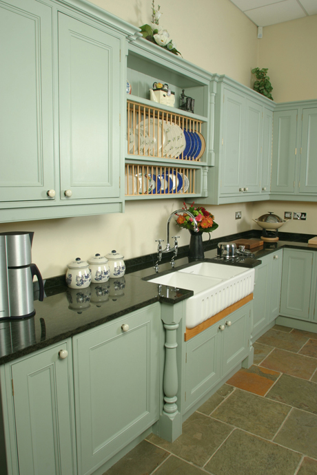 Landhausstil-Küche mit grünen Schränken, Keramikspüle und Arbeitsplatte aus Granit Gemütliche Küche im Landhausstil mit grünen Schränken, Keramikspüle, schwarzen Granit-Arbeitsflächen, Keramikdosen und einem Tellerregal über der Spüle