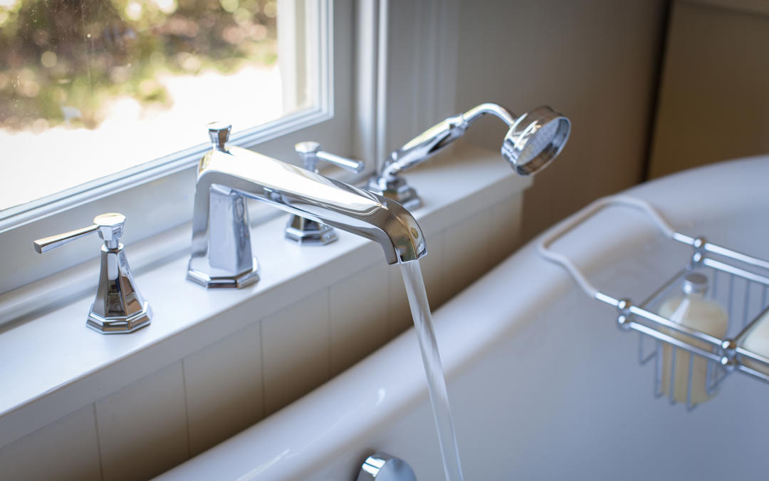 Moderne Chrom-Badewannenarmatur mit zwei Griffen und Handbrause; Wasser läuft in eine freistehende Wanne – hochwertige Wannenrandarmatur fürs Bad