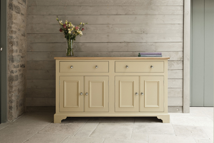 Landhausstil Anrichte aus Holz mit 4 Türen und 2 Schubladen, cremefarben, dekoriert mit Vase und Büchern Cremefarbene Anrichte im Landhausstil aus Holz, mit vier Türen und zwei Schubladen, darauf steht eine Vase mit Blumen und einige Bücher.