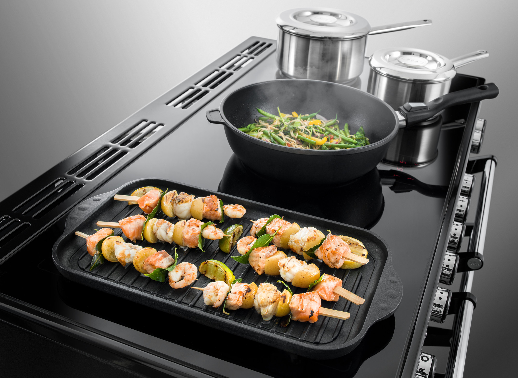 Induktionskochfeld mit Grillplatte, Fisch- und Gemüsespießen sowie Pfanne und Töpfen in moderner Küche Induktionskochfeld mit Grillplatte, darauf Spieße mit Lachs, Garnelen, Kartoffeln und Gemüse, daneben Pfanne mit grünem Gemüse und zwei Edelstahltöpfe
