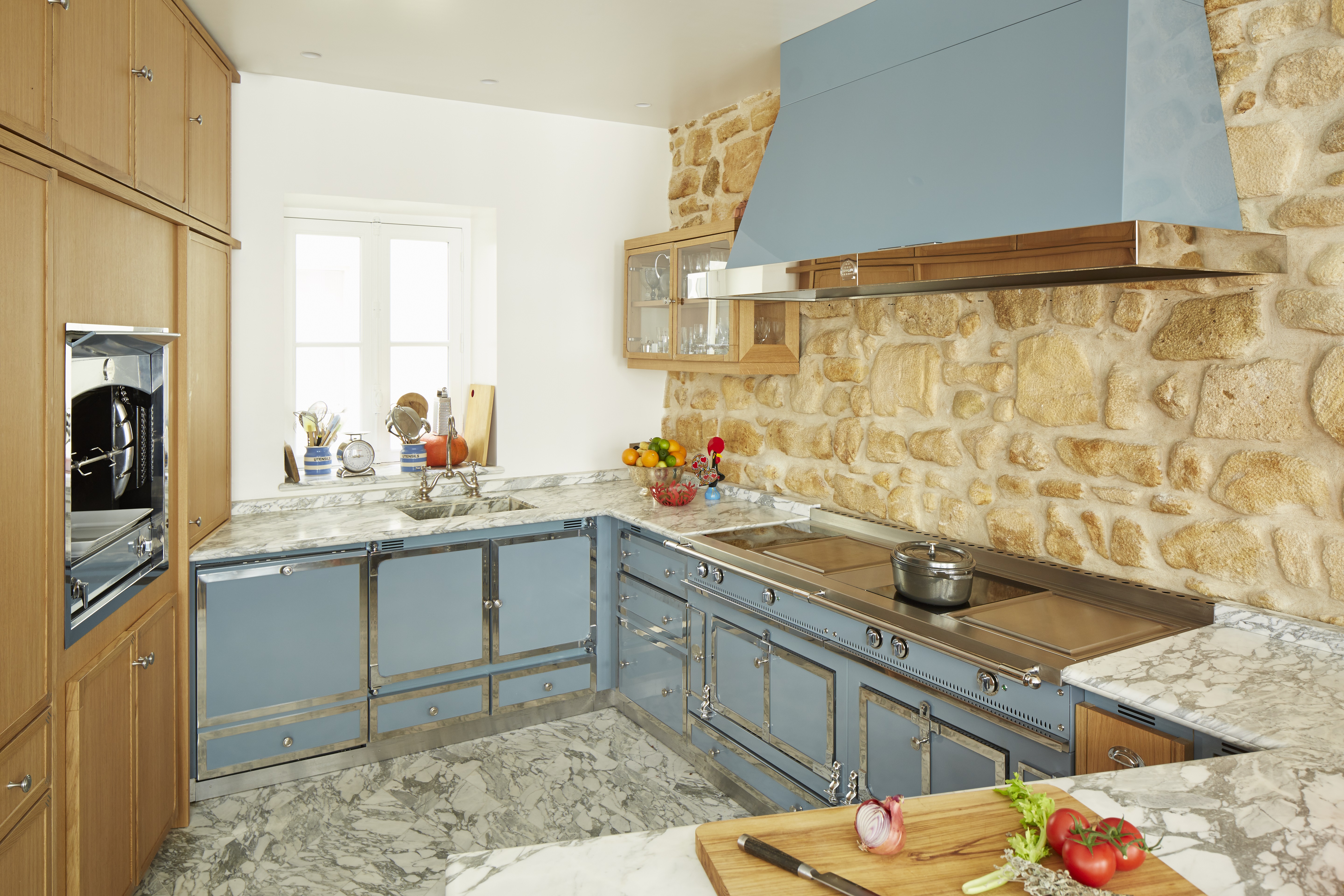 Moderne Landhausküche mit blauen Küchenmöbeln, Steinwand und Marmorarbeitsfläche Helle Küche mit blauen Unterschränken, Marmorarbeitsfläche, Steinwand, Einbauofen und frischem Gemüse auf dem Schneidebrett