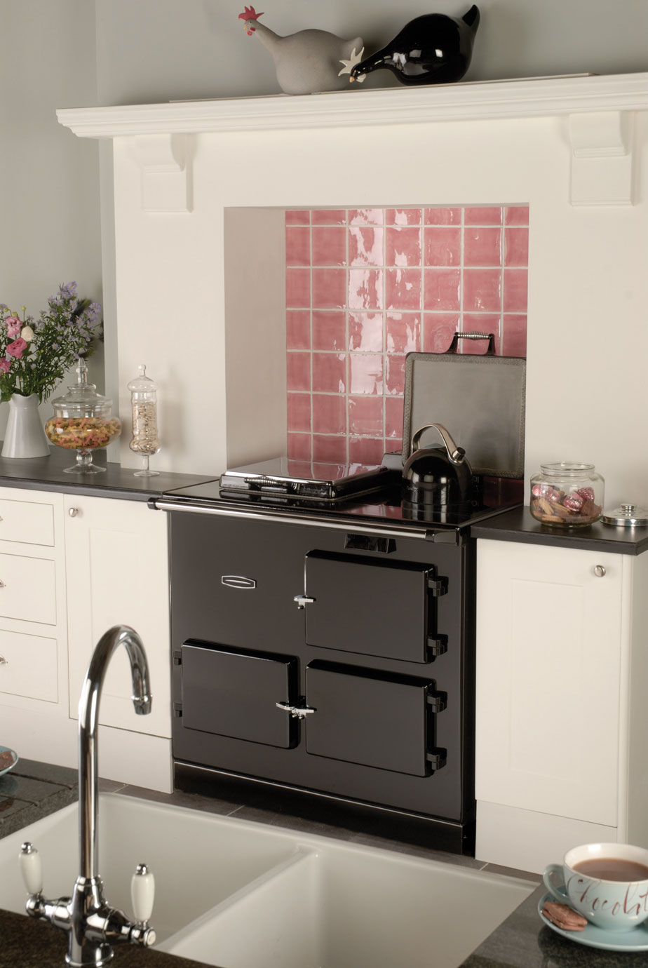 Schwarzer Retro-Ofen in moderner Küche mit rosa Fliesenspiegel und Dekoration Moderner Küchenbereich mit schwarzem Retro-Ofen, rosa Fliesenspiegel, Blumen und Keramikfigur auf dem Kaminsims