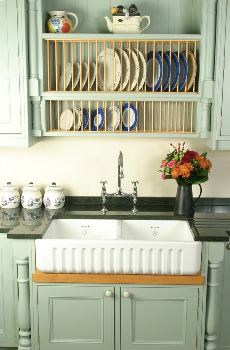 Landhausküche mit Doppelspülbecken, Tellerregal und Blumenvase auf schwarzer Arbeitsplatte Küchenspüle im Landhausstil mit grünlichen Schränken, Tellerregal mit Geschirr und Blumenvase auf der Arbeitsplatte