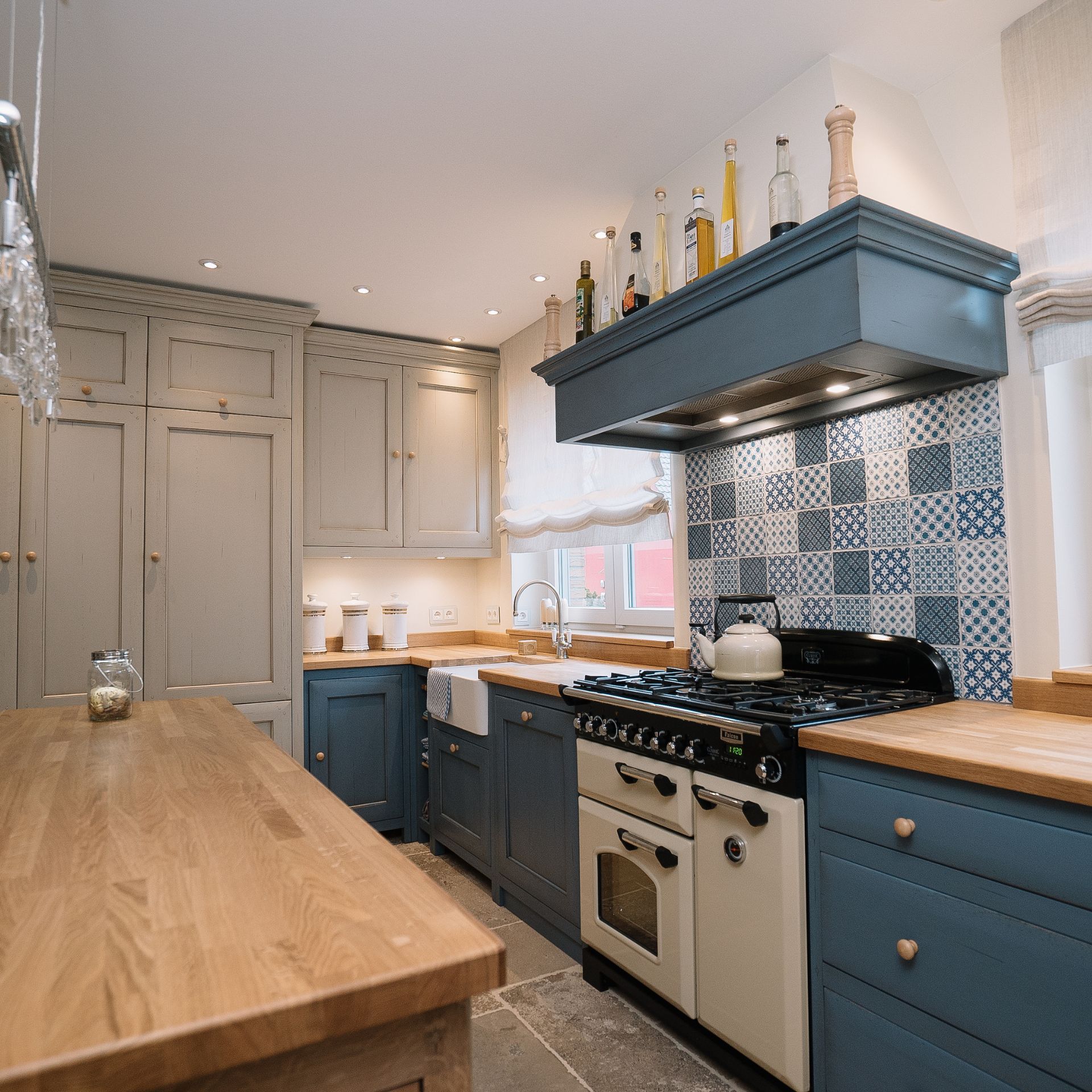 Gemütliche Landhausküche mit blauen Unterschränken, Holz-Arbeitsplatten, cremefarbenem Gasherd mit Backofen, blauer Fliesenrückwand, Keramikspüle am Fenster und Kücheninsel aus Holz