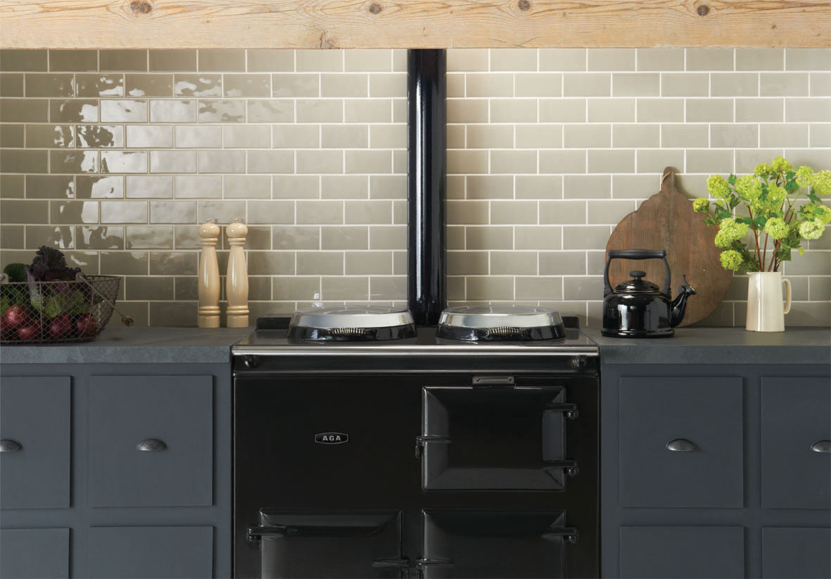 Klassischer schwarzer AGA-Herd in moderner Landhausküche mit Fliesenspiegel Ein schwarzer AGA-Herd steht zwischen grauen Schränken, davor Gemüsekorb, Salz- und Pfeffermühle sowie eine schwarze Teekanne und Blumen auf der Arbeitsfläche.