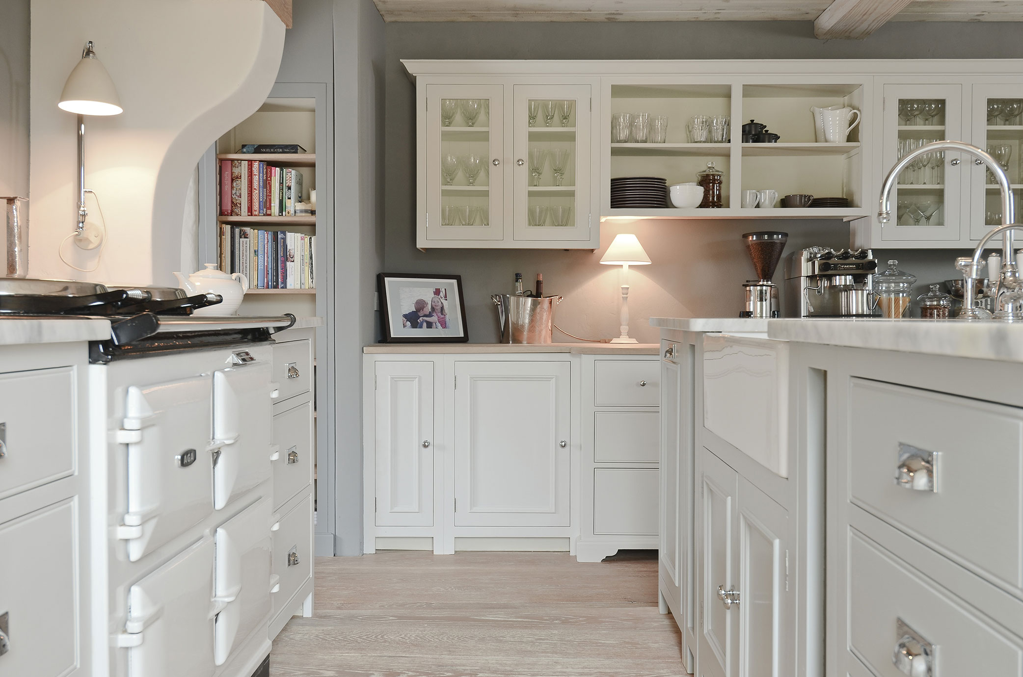 Moderne Landhausküche mit weißen Schränken, Kücheninsel und stilvoller Einrichtung Helle Landhausküche mit weißen Fronten, Glastüren, Kücheninsel, Herd und stilvollen Accessoires wie Lampen, Kaffeemaschine und Bücherregal