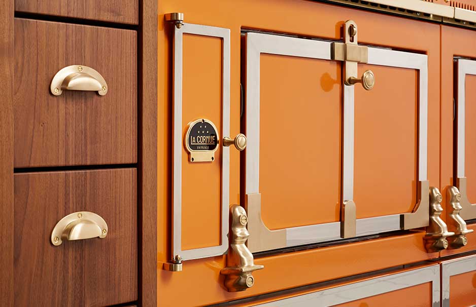 Detailansicht eines hochwertigen, orangefarbenen La Cornue Herdes mit goldfarbenen Griffen und Holzschubladen Hochwertiger, orangefarbener La Cornue Herd mit goldfarbenen Griffen und einem Teil von Holzschubladen im klassischen Design