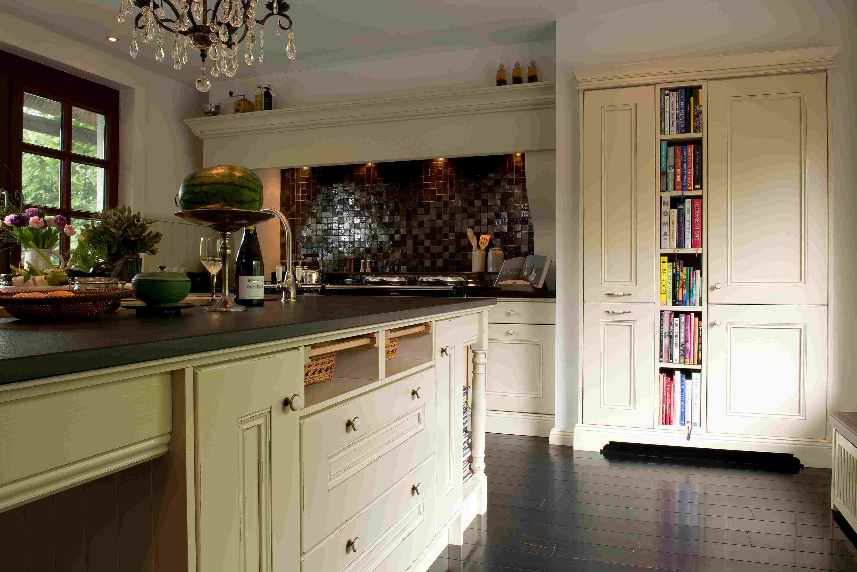 Moderne Landhausküche mit cremefarbenen Schränken, Kochinsel und integriertem Bücherregal Gemütliche Küche im Landhausstil mit heller Einrichtung, dunkler Arbeitsplatte, Kochinsel und offenem Regal für Kochbücher