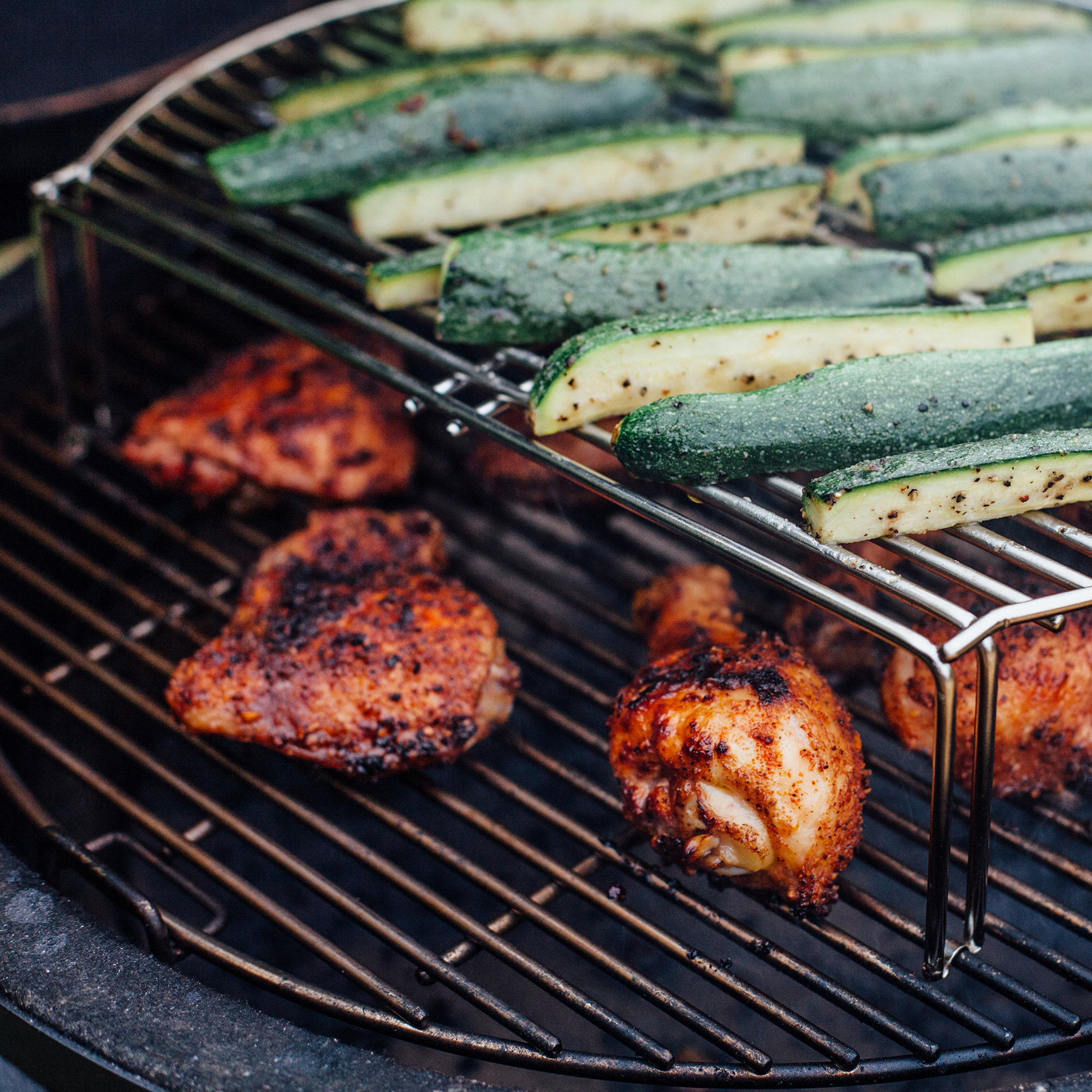 Gegrillte Hähnchenschenkel und Zucchini auf doppeltem Grillrost, frisch gewürzt und knusprig zubereitet Frisch gewürzte Hähnchenschenkel und Zucchinistreifen auf einem Grill mit doppeltem Rost beim Grillen