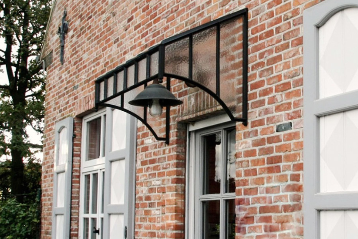Vordach aus Glas und Metall mit integrierter Lampe über einer Haustür an einem Backsteingebäude Modernes Glas- und Metallvordach mit schwarzem Rahmen und integrierter Außenlampe, montiert über einer Haustür an einer Fassade aus roten Ziegelsteinen