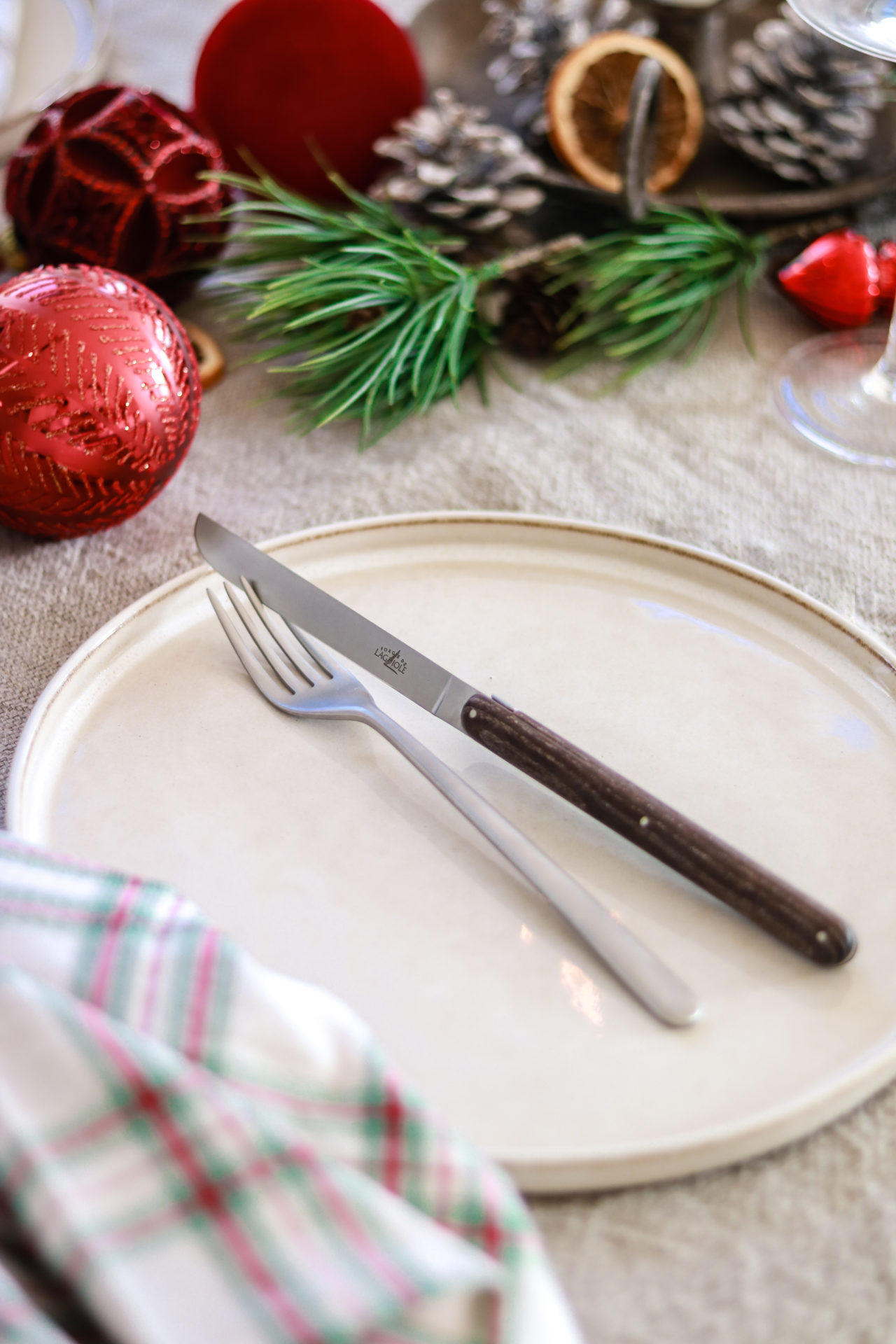 Weihnachtlich gedeckter Tisch mit Besteck und Dekoration Weißer Teller mit Messer und Gabel, festlich gedeckt mit roter Weihnachtsdeko, Kieferzweigen und Zapfen
