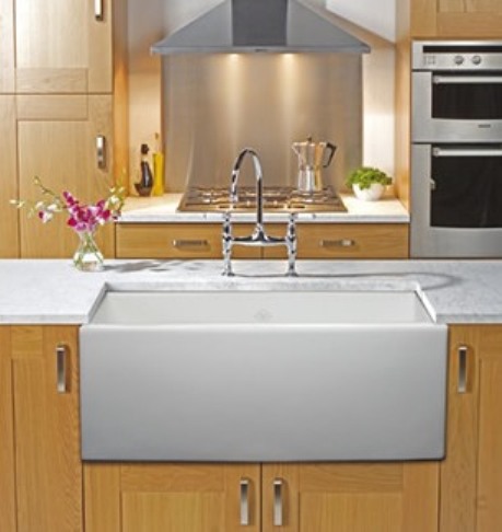 Moderne Küche mit großer Keramik-Spüle, Edelstahl-Armatur, Holzschränken und Einbaugeräten Helle, moderne Küche mit großer, weißer Keramik-Spüle, zentral platzierter Edelstahl-Armatur und hellen Holzunterschränken. Im Hintergrund befinden sich ein Herd mit Dunstabzugshaube, ein Backofen und eine Kaffeemaschine. Links steht eine Glasvase mit Blu