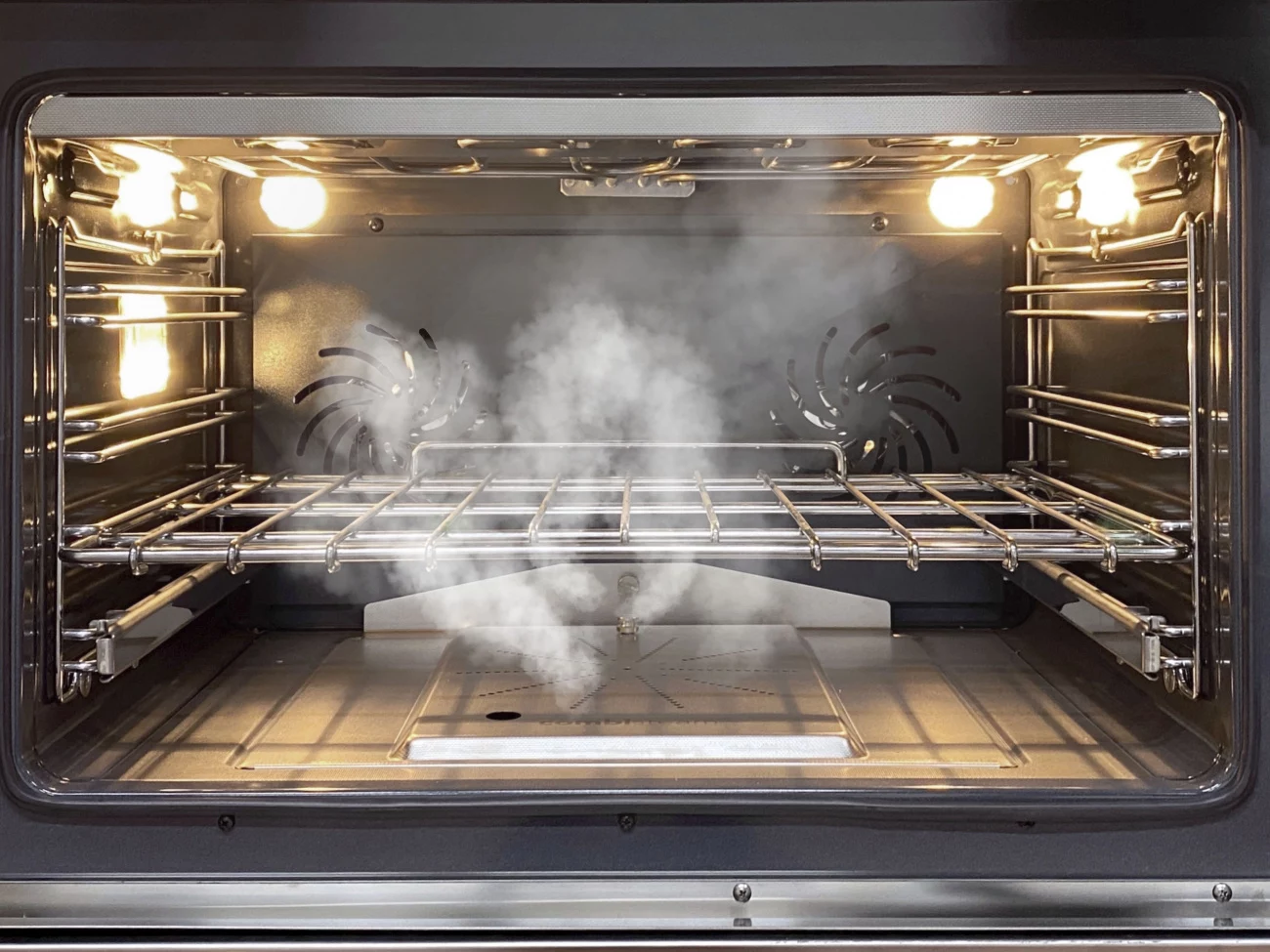 Beleuchteter Backofen-Innenraum mit aufsteigendem Dampf, Edelstahlrost und Umluftventilatoren – ideal für Dampf-Reinigung oder schonendes Garen