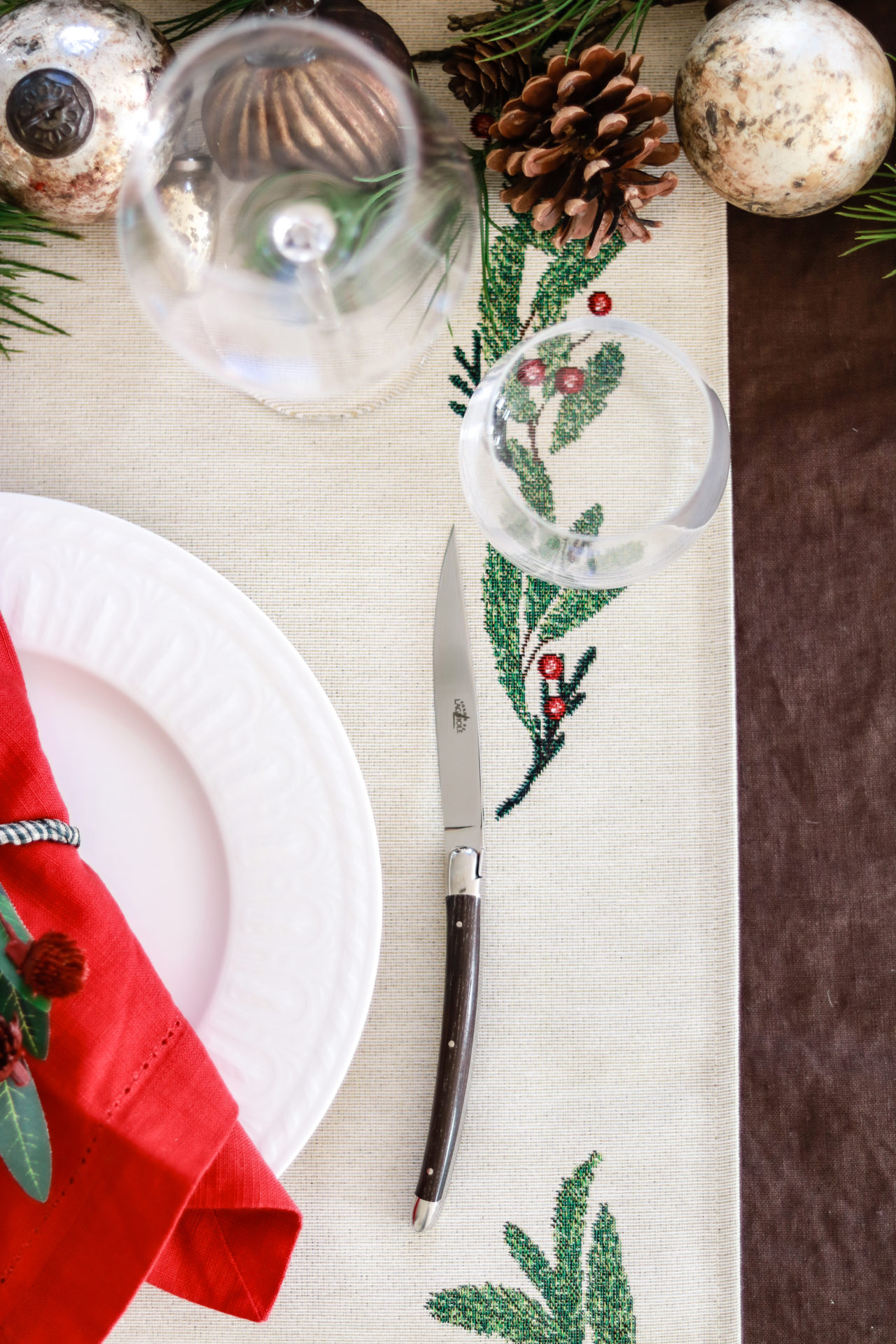 Festlich gedeckter Tisch mit Weihnachtsdeko, weißem Teller, roter Serviette und Messer Weihnachtlich dekorierter Tisch mit Keramik-Teller, roten Servietten, Glas, Messer, Tannenzapfen und besticktem Tischläufer mit Zweigen