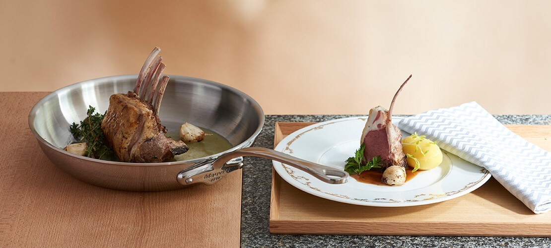 Edelstahlpfanne mit Lammkarree und angerichtetem Lammgericht auf edlem Porzellanteller Lammkarree in Edelstahlpfanne mit Kräutern und Knoblauch, daneben ein angerichtetes Lammgericht mit Sauce und Kartoffelpüree auf einem Porzellanteller mit Stoffserviette