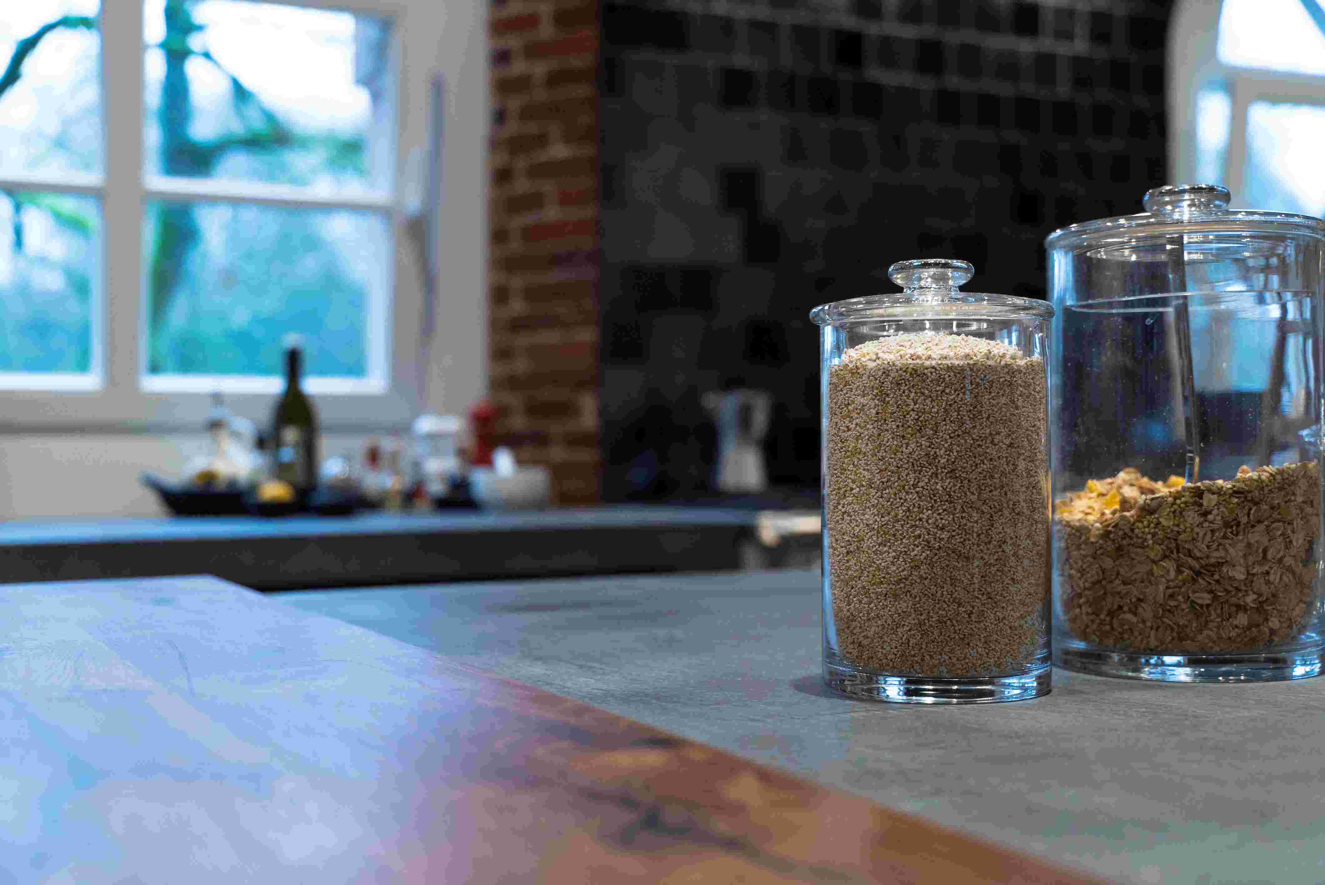 Zwei Glasgefäße, eines gefüllt mit Getreide und das andere mit Müsli, stehen auf einer Küchenarbeitsplatte vor einem Fenster mit Tageslicht.