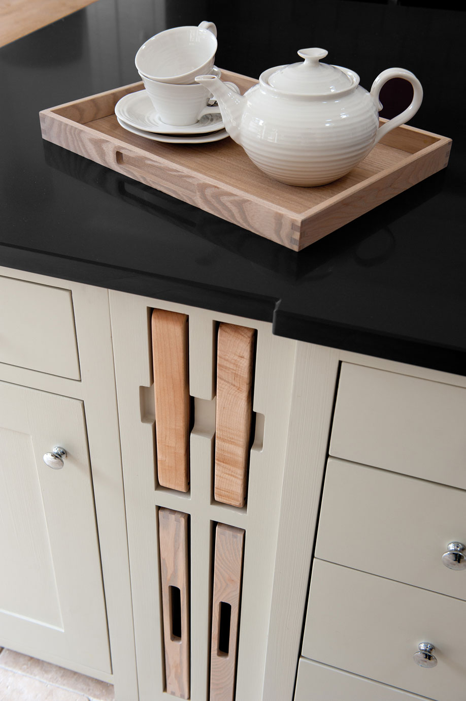 Schwarze Küchenarbeitsplatte mit Holztablett und weißem Teeservice sowie integrierten Holzschneidebrettern Modernes Küchendesign mit schwarzer Arbeitsplatte, Holztablett mit weißer Teekanne und Tassen sowie integrierten Holzschneidebrettern in einem hellen Unterschrank