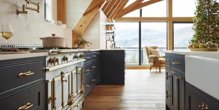 Hochwertige Landhausküche in Dunkelblau mit Messingbeschlägen, großer Range mit Backofen, Farmhouse-Keramikspüle und Holzboden; bodentiefe Fenster mit Bergblick, festlich dekoriert