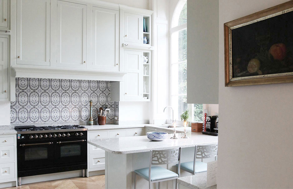 Moderne weiße Küche mit Kücheninsel, großem Fenster und gemusterter Fliesenrückwand Helle, moderne Küche mit weißen Schränken, einer Kücheninsel, Barhockern, großem Fenster, gemusterten Fliesen und einem Backofen