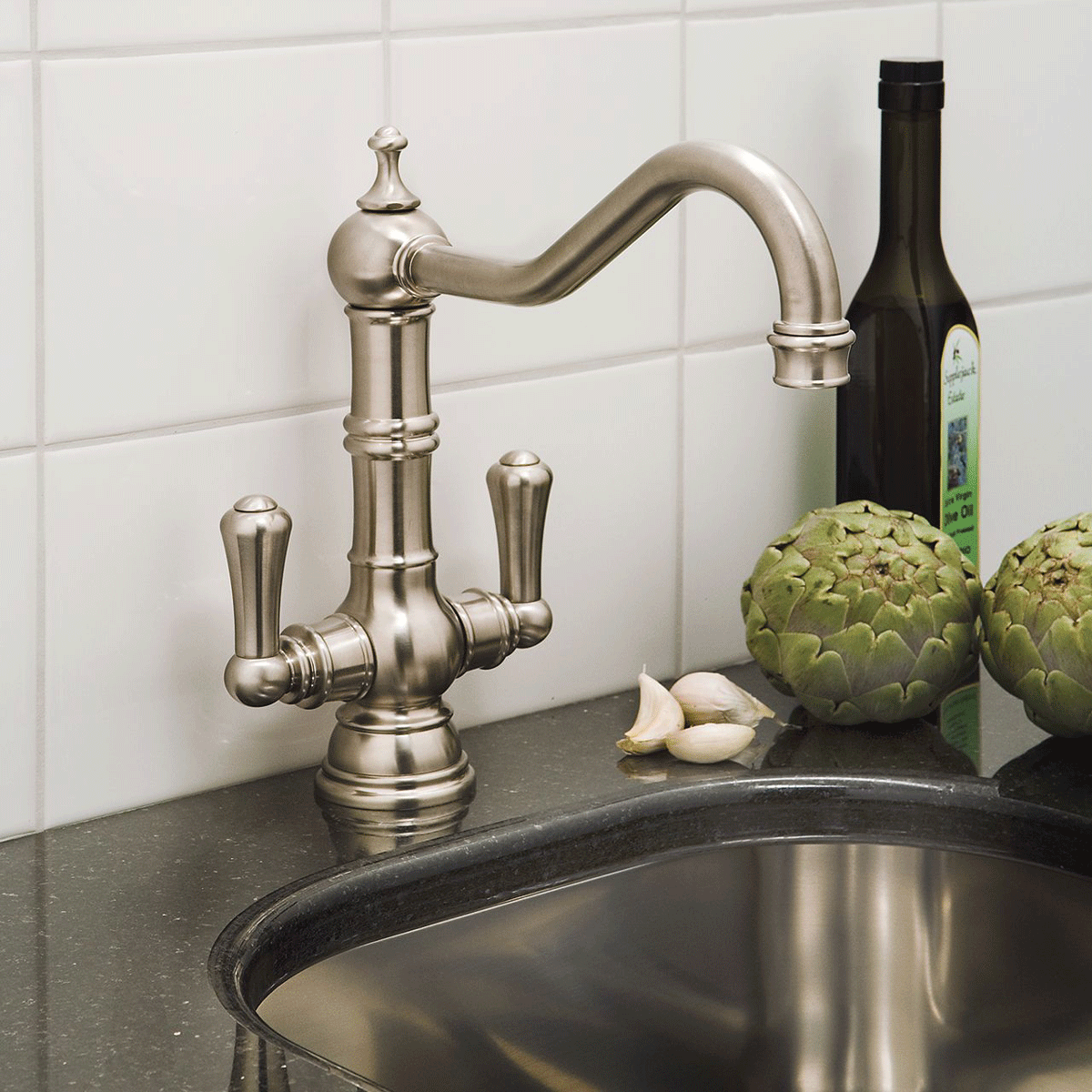 Elegante Küchenarmatur im Retro-Design aus Edelstahl mit zwei Griffen, montiert auf schwarzer Arbeitsplatte Küchenarmatur aus Edelstahl im klassischen Design neben Spüle, Artischocken, Knoblauch und Olivenölflasche auf schwarzer Arbeitsplatte