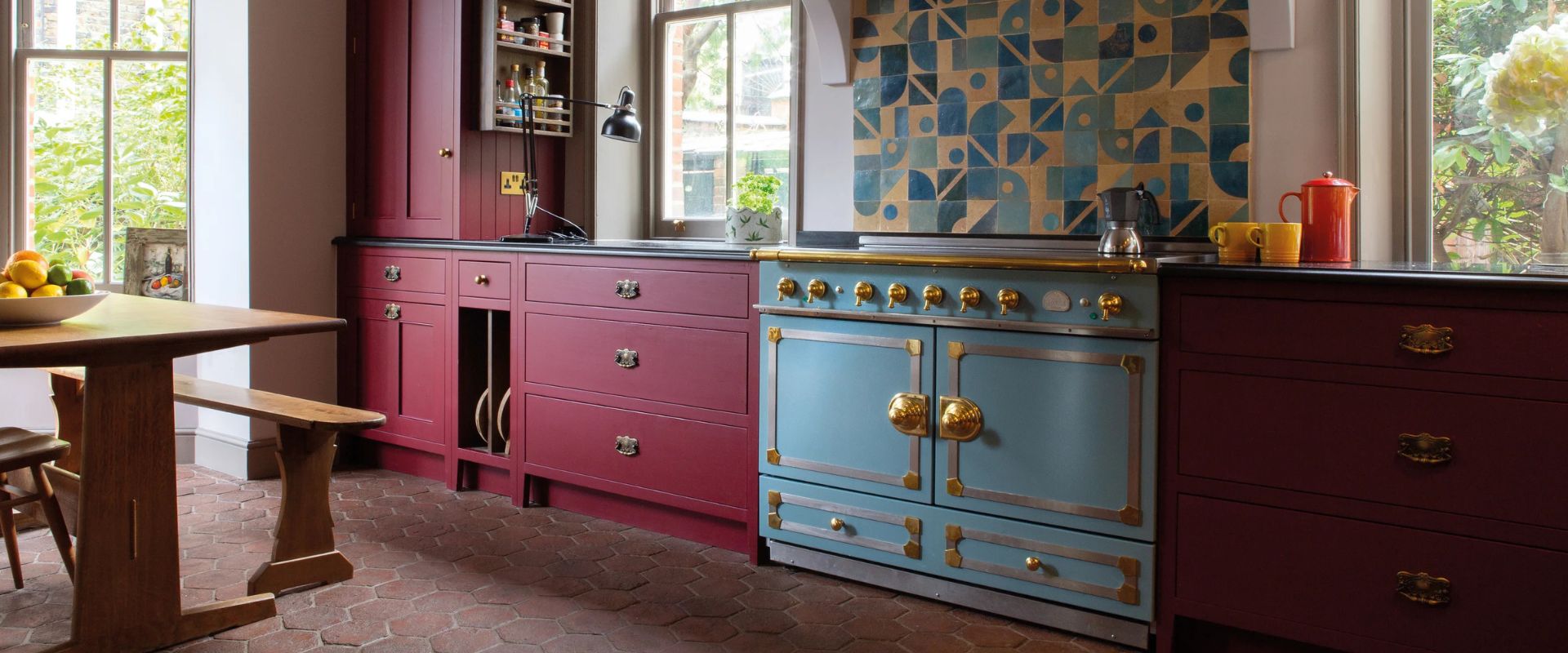 Blauer Standherd mit Messingknöpfen in weinroter Landhausküche mit geometrischer Fliesenrückwand; hochwertiger Range Cooker als Blickfang.