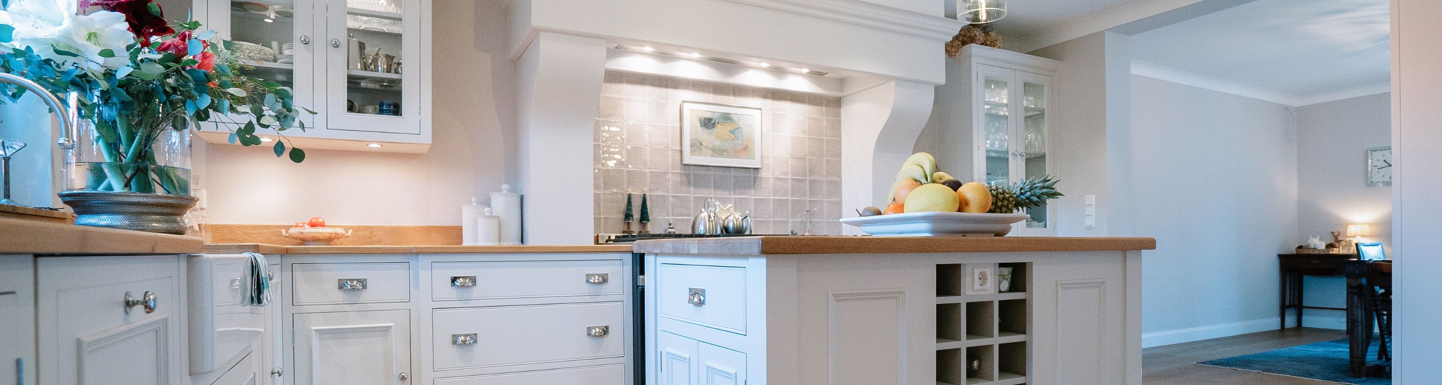 Helle Landhausküche mit weißen Schränken, Holzarbeitsplatte, Kücheninsel und Obstschale, grauem Fliesenspiegel, Unterbauleuchten sowie angrenzendem Wohnbereich mit Schreibtisch