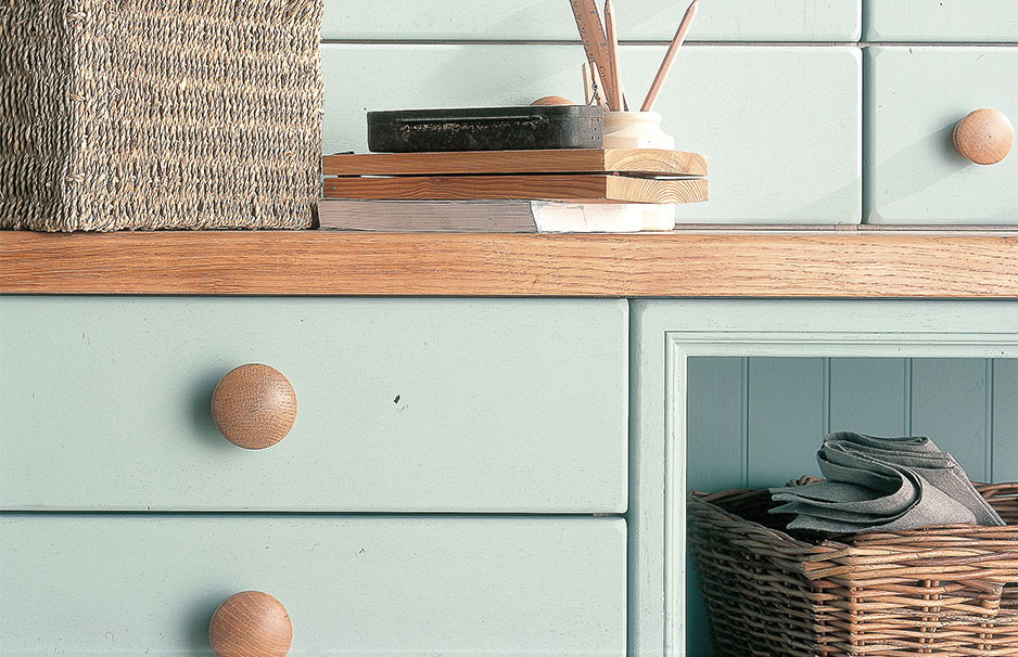Detailansicht einer mintgrünen Kommode mit Holzakzenten, Körben und Dekoration Mintgrüne Kommode mit Holzgriffen, geflochtenem Korb und Büchern auf der Ablage, sowie gefalteten Tüchern im Korb