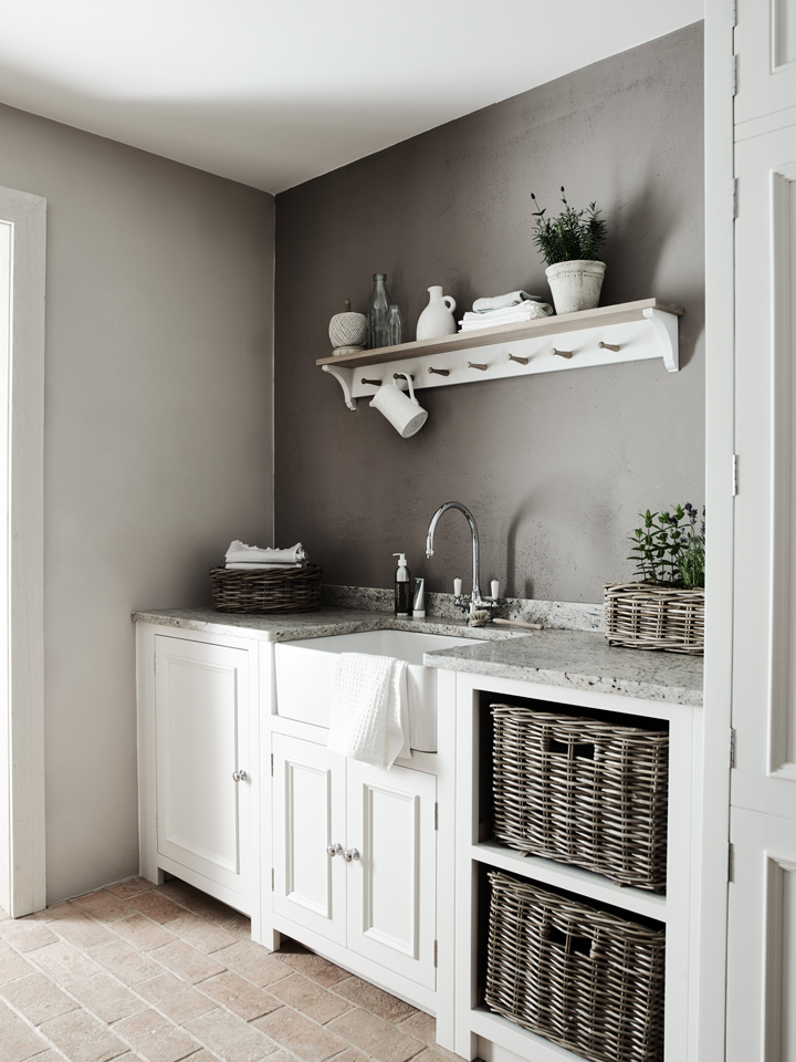 Landhausstil-Waschküche mit weißen Schränken, Naturstein-Arbeitsplatte und geflochtenen Aufbewahrungskörben Helle Waschküche im Landhausstil mit weißen Unterschränken, Keramikspüle, Naturstein-Arbeitsfläche, offenen Regalen und geflochtenen Körben als Stauraum