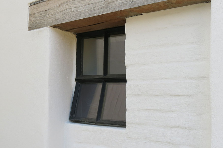 Kleines rechteckiges Fenster mit schwarzem Rahmen in einer weißen Wand Schräg eingebautes, kleines Fenster mit schwarzem Rahmen in einer weiß verputzten Hauswand mit Holzbalken darüber