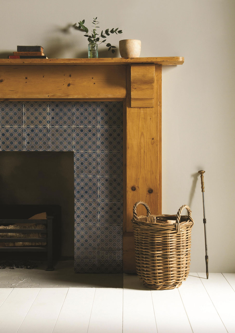 Kaminverkleidung aus Holz mit dekorativen blauen Fliesen und Korb für Kaminholz Gemütlicher Kamin mit Holzverkleidung, dekorativen blauen Fliesen, einem Weidenkorb für Kaminholz und Kaminbesteck