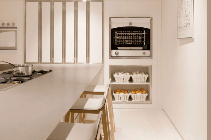 Moderne weiße Küche mit Gasherd, Sitzbereich und Regalen mit Obstkörben Helle, minimalistische Küche mit Gasherd, Backofen, weißen Stühlen und mehreren Körben mit Eiern und Orangen in einem Regal