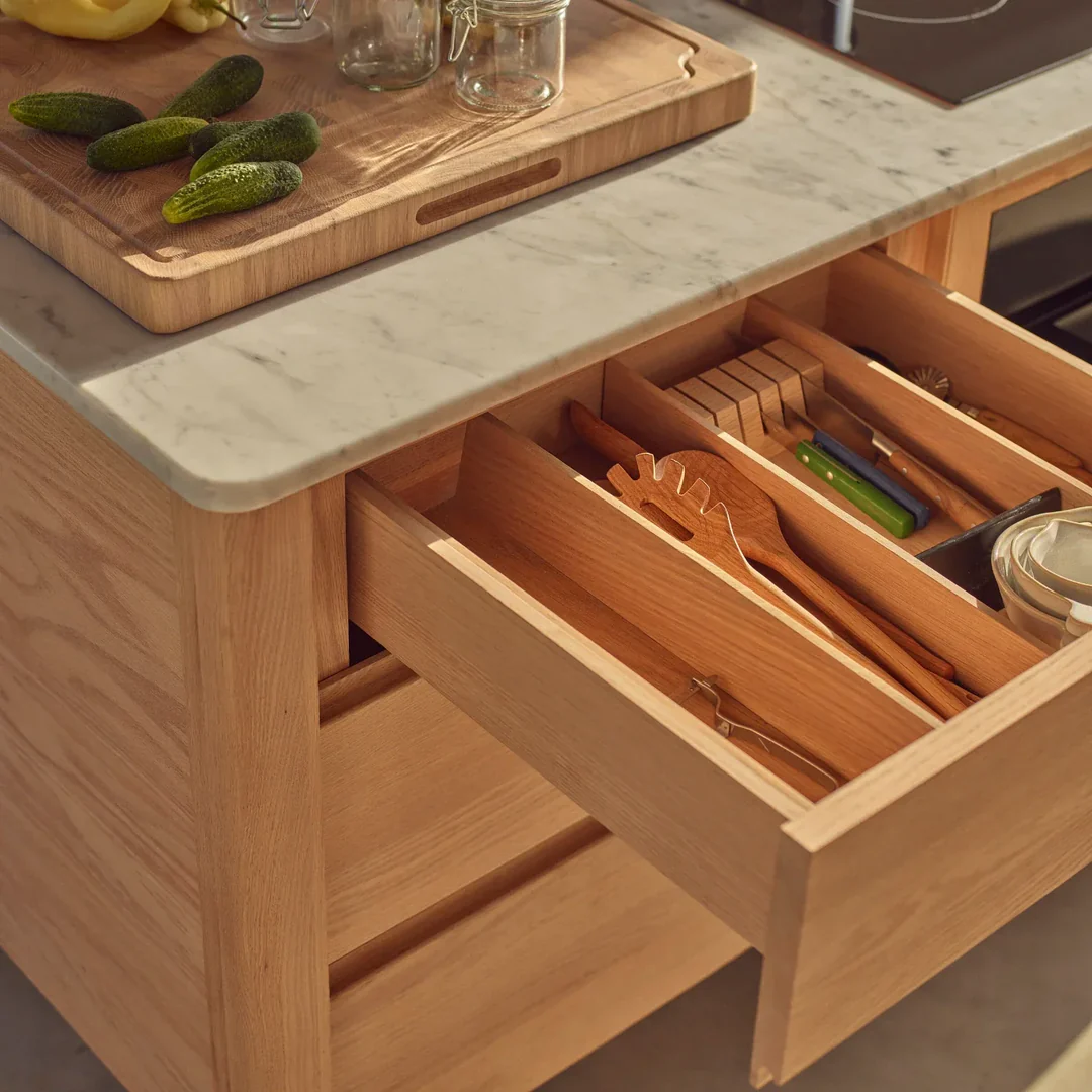 Geöffnete Schublade mit Holz-Besteckorganizer in Kücheninsel aus Eiche; darauf Marmor-Arbeitsplatte mit Schneidebrett, Gurken und Gläsern – ordentliche Aufbewahrungslösung für die Küche
