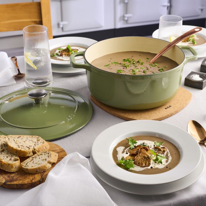 Eleganter Esstisch mit grüner Gusseisen-Kasserolle, servierter Pilzsuppe und frischem Brot Gedeckter Tisch mit grüner Gusseisen-Kasserolle, cremiger Pilzsuppe in Tellern, frischem Brot und Wassergläsern