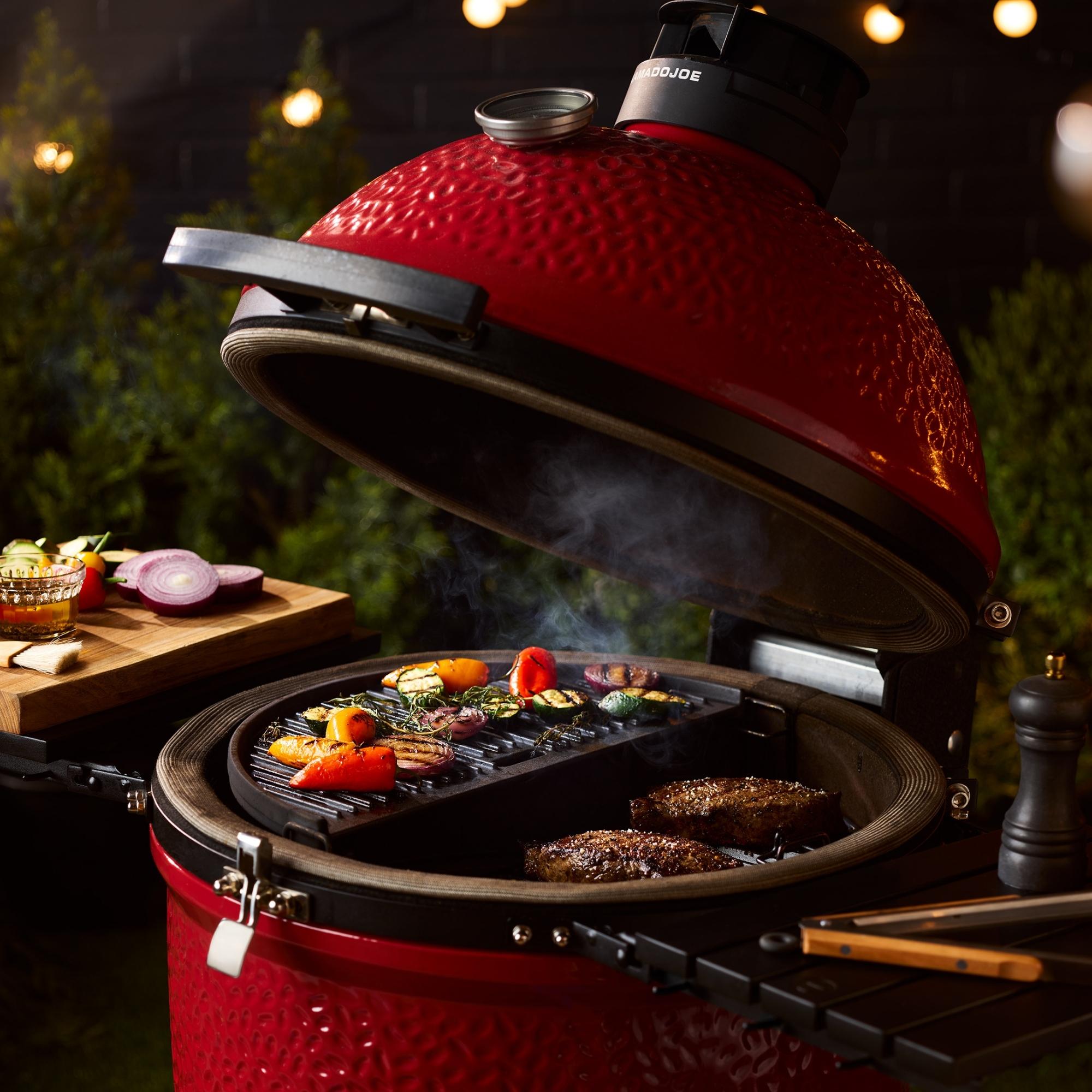 Kamado Joe Keramikgrill in Rot mit geöffnetem Deckel und Grillgut – Perfekt für BBQ, Steak und Gemüsegrillen im Garten Roter Kamado Joe Keramikgrill mit offenem Deckel, Grillrost mit gegrilltem Fleisch und buntem Gemüse, Holztisch mit Zwiebelscheiben und Kräutern im Hintergrund, stimmungsvolle Gartenbeleuchtung