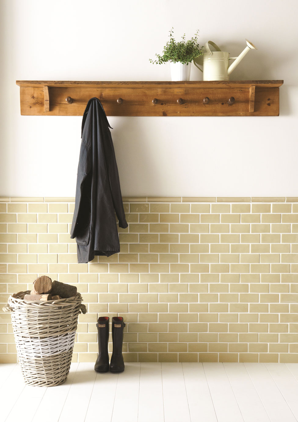 Flur mit Wandregal aus Holz, Haken, Gummistiefeln, Wäschekorb mit Holzscheiten und Dekoration Wandregal aus Holz mit Haken, daran eine Jacke, oben Blumentopf und Gießkanne, darunter Korb mit Holz und Gummistiefel auf weißem Boden