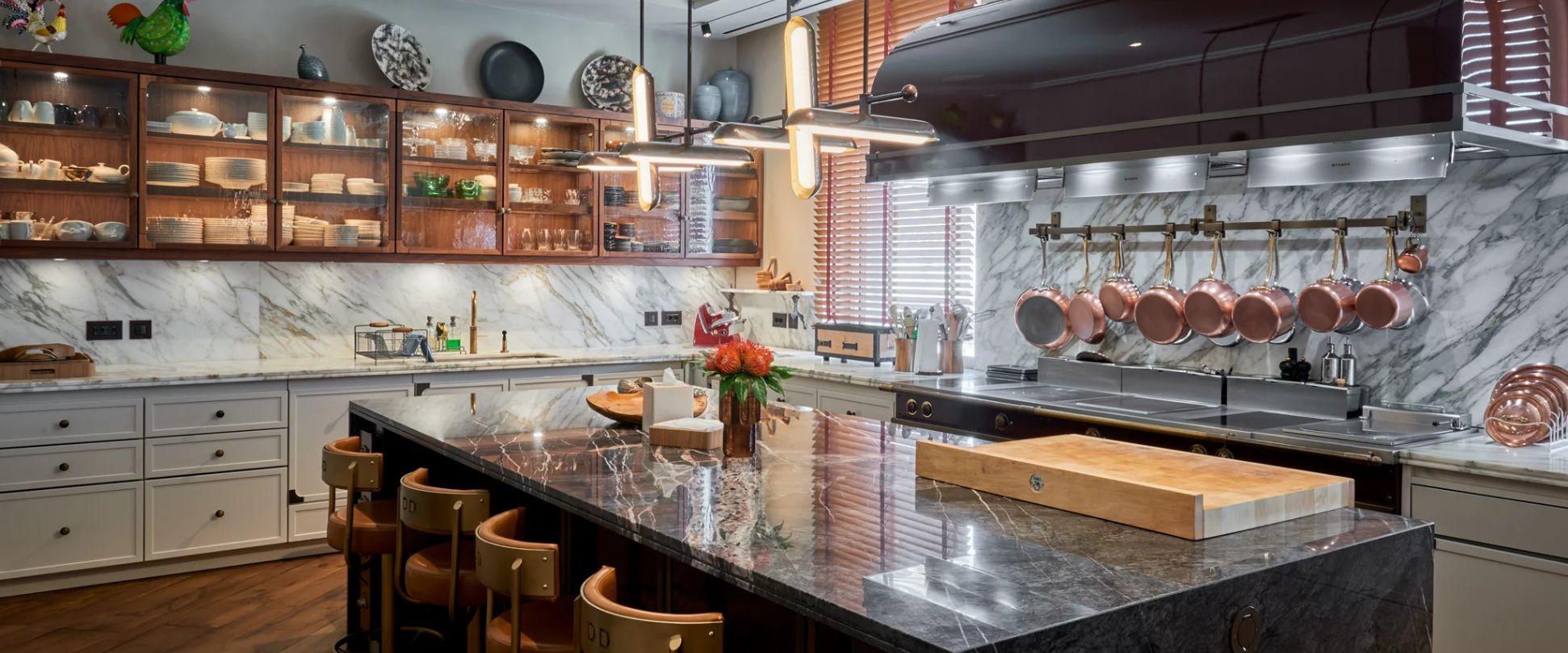Hochwertige Designküche mit großer Kücheninsel aus dunklem Marmor, marmorierter Rückwand, Hängereihe aus Kupfertöpfen, beleuchteten Vitrinenschränken und professioneller Kochzone – elegantes Premium-Küchenambiente