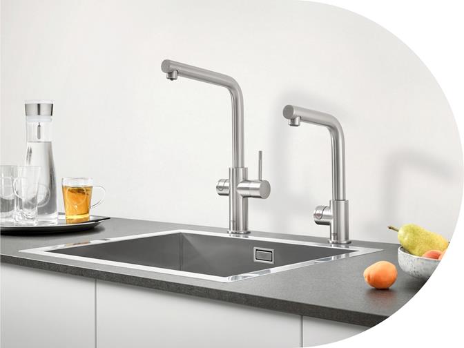 Moderne Edelstahl-Küchenspüle mit zwei Armaturen auf dunkler Arbeitsplatte Küchenspüle mit zwei eleganten Edelstahl-Armaturen, daneben Obstschale und Gläser auf der Arbeitsplatte